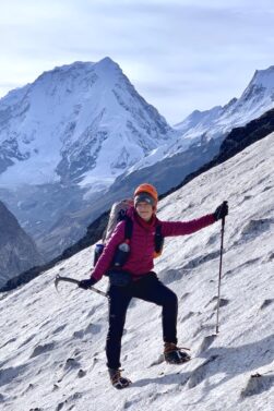 Mathilde Godbout, en tenue d'alpiniste, escalade une pente enneigée sur l'Himalaya à l'aide d'un piolet et d'un bâton de trekking. En arrière-plan, on aperçoit des montagnes enneigées et un ciel dégagé. - Karavaniers