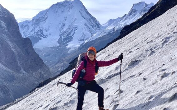 Mathilde Godbout, en tenue d'alpiniste, escalade une pente enneigée sur l'Himalaya à l'aide d'un piolet et d'un bâton de trekking. En arrière-plan, on aperçoit des montagnes enneigées et un ciel dégagé. - Karavaniers