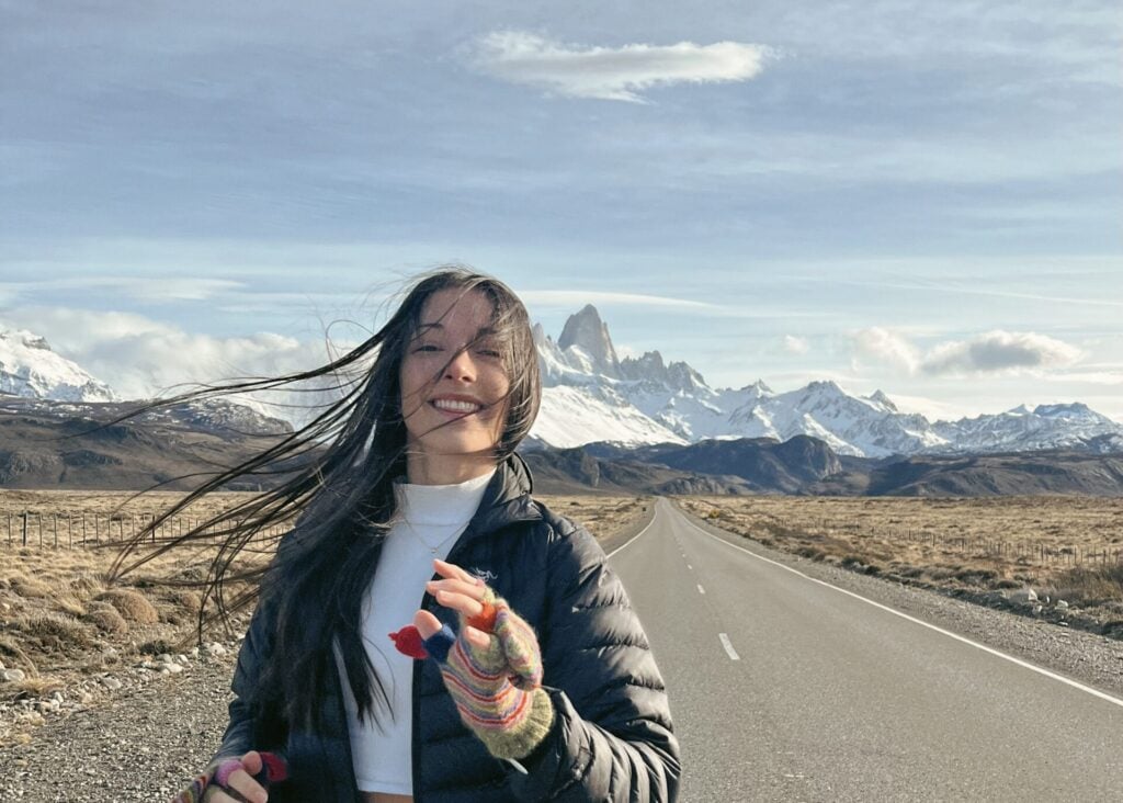 Mathilde Godbout sourit sur une route déserte, le vent soufflant dans ses cheveux. Elle porte une veste noire et des gants colorés, tandis que des montagnes enneigées s'élèvent à l'arrière-plan sous un ciel partiellement nuageux. - Karavaniers