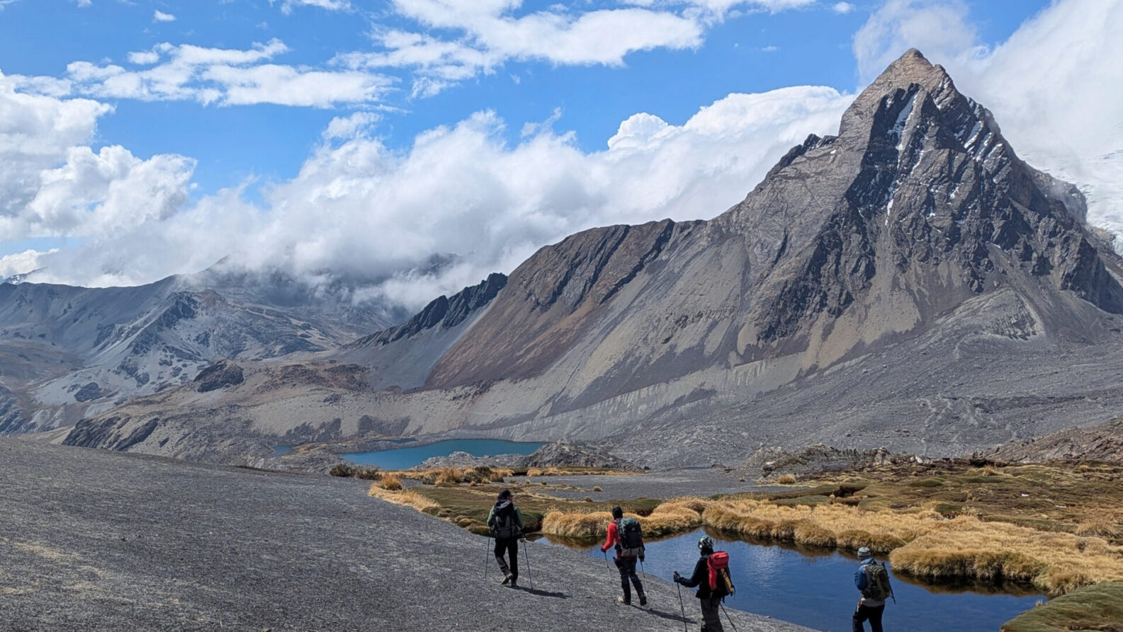 Quatre randonneurs avec des sacs à dos marchent sur un sentier au bord d'un petit lac dans les montagnes de Bolivie, avec des pentes rocheuses et un sommet enneigé en arrière-plan sous un ciel partiellement nuageux. - Karavaniers