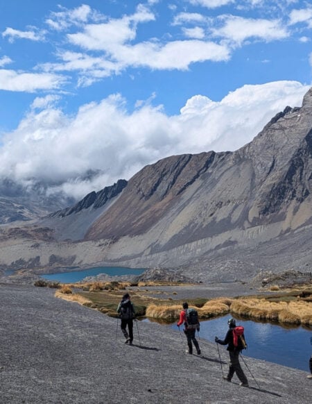 Quatre randonneurs avec des sacs à dos marchent sur un sentier au bord d'un petit lac dans les montagnes de Bolivie, avec des pentes rocheuses et un sommet enneigé en arrière-plan sous un ciel partiellement nuageux. - Karavaniers