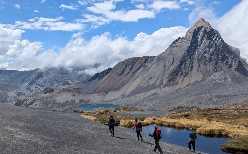 Quatre randonneurs avec des sacs à dos marchent sur un sentier au bord d'un petit lac dans les montagnes de Bolivie, avec des pentes rocheuses et un sommet enneigé en arrière-plan sous un ciel partiellement nuageux. - Karavaniers