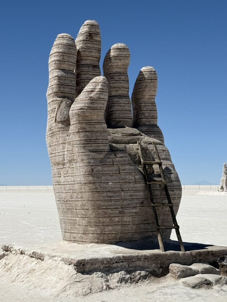 Une grande sculpture en pierre représentant une main humaine droite se dresse dans un désert plat et aride, sous un ciel bleu clair. Une échelle en bois s'appuie sur la main, s'élevant vers les doigts. - Karavaniers