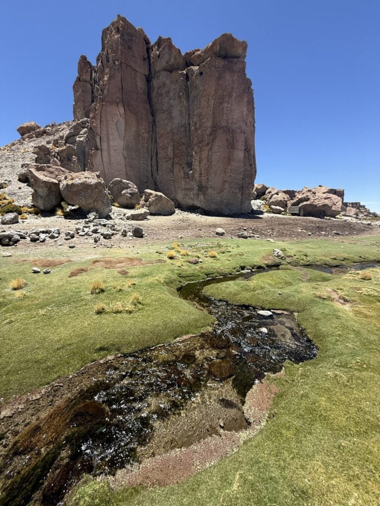 Une haute formation rocheuse s'élève dans une zone sèche et rocailleuse. Un ruisseau étroit et peu profond coule dans l'herbe verte au premier plan, sous un ciel bleu clair. - Karavaniers