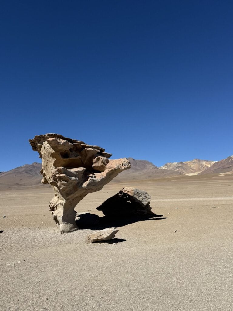 Une grande formation rocheuse de forme unique se dresse sur une plaine désertique plate et sablonneuse, avec des montagnes en arrière-plan, sous un ciel bleu clair. - Karavaniers