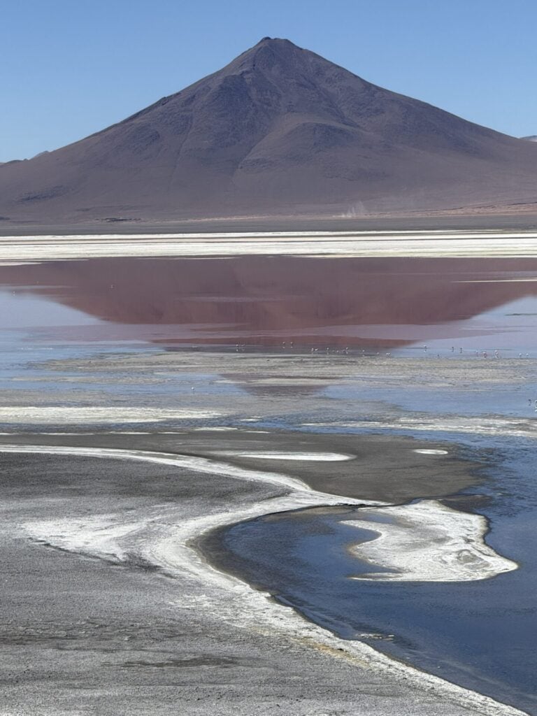 Une montagne sombre se dresse derrière un lac réfléchissant et peu profond, avec des dépôts blancs salés sur le rivage. Le reflet de la montagne est visible dans l'eau calme sous un ciel bleu clair. - Karavaniers