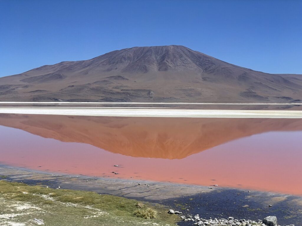 Une montagne se reflète dans un lac rouge rosé avec une bande de rive blanche. Le premier plan montre un terrain rocailleux et herbeux sous un ciel bleu clair. - Karavaniers