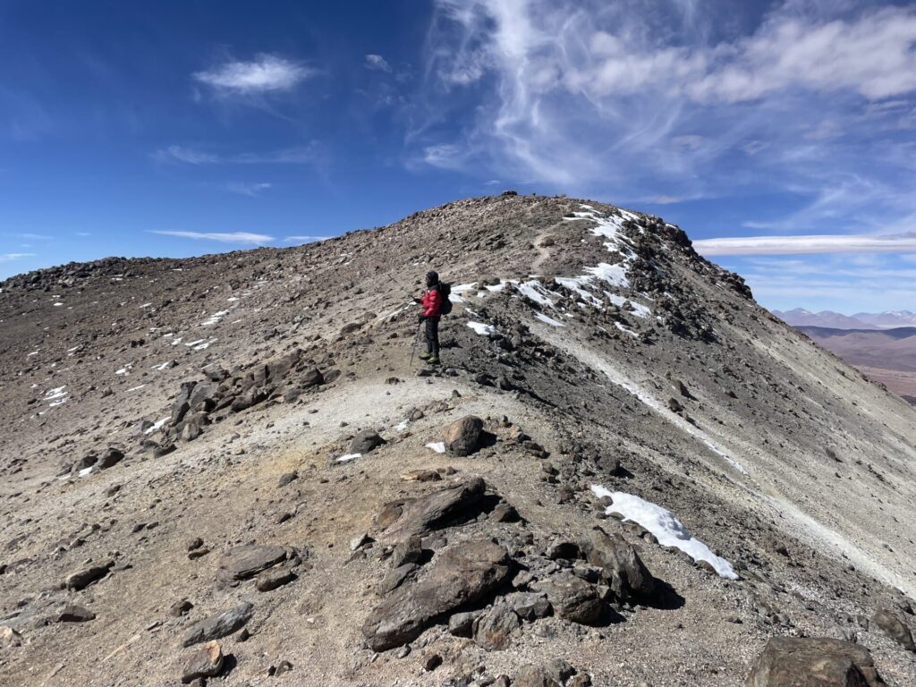 Une personne portant une veste rouge se tient sur la crête d'une montagne rocheuse avec des plaques de neige sous un ciel bleu avec des nuages épars. - Karavaniers