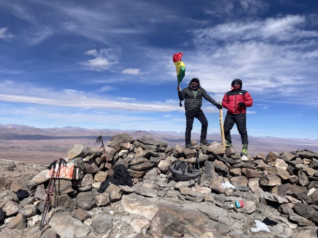 Deux personnes en tenue de plein air se tiennent sur un sommet rocheux. L'une d'elles tient un drapeau coloré. Un sac à dos et des bâtons de randonnée sont posés sur les rochers. En arrière-plan, on aperçoit des montagnes et un ciel large et dégagé. - Karavaniers