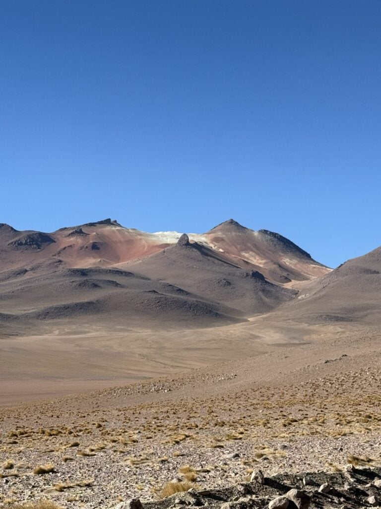 Terrain aride et rocailleux avec une végétation clairsemée au premier plan, menant à une chaîne de montagnes brunes et rougeâtres sous un ciel bleu clair. - Karavaniers