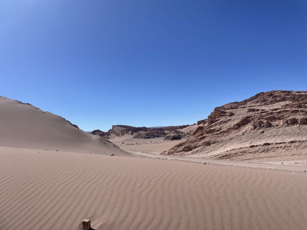 Un paysage désertique sablonneux avec des dunes ondulées au premier plan, des collines rocheuses à l'arrière-plan et un ciel bleu clair au-dessus de la tête. - Karavaniers