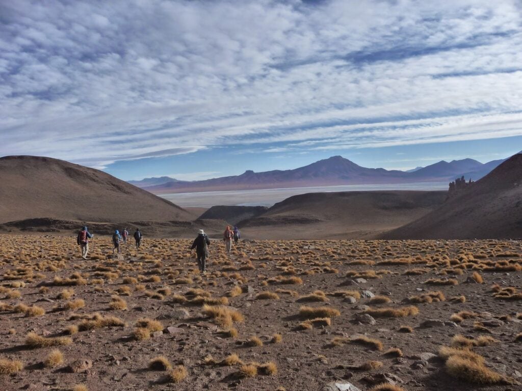 Un groupe de personnes marche dans un paysage sec et rocailleux avec de l'herbe jaune éparse, entouré de collines. Des montagnes et un lac sont visibles au loin dans un ciel parsemé de nuages. - Karavaniers
