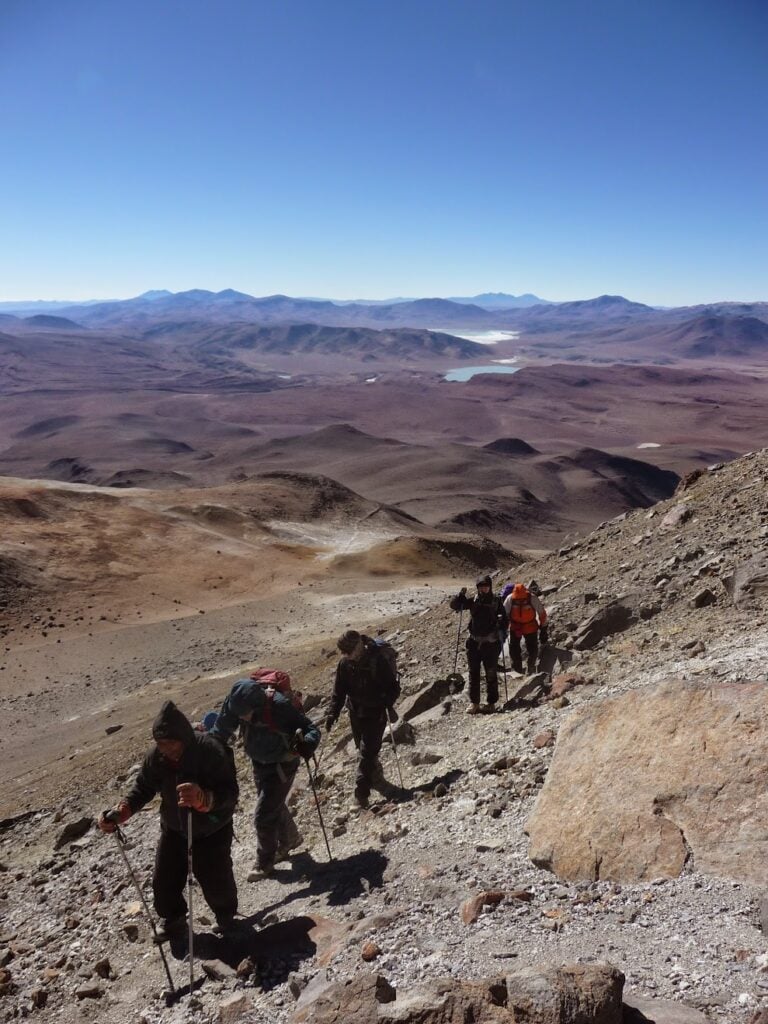 Un groupe de randonneurs munis de sacs à dos et de bâtons de trekking gravit un versant rocheux d'une montagne sous un ciel bleu clair, avec en arrière-plan un vaste paysage aride et des lacs lointains. - Karavaniers