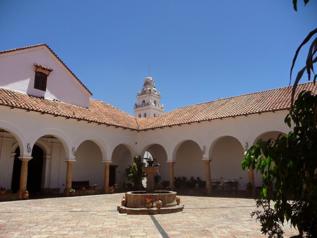 Une cour avec des allées voûtées, une fontaine centrale en pierre et des toits en tuiles de terre cuite. À l'arrière-plan, une tour d'horloge blanche s'élève au-dessus des bâtiments sous un ciel bleu clair. - Karavaniers