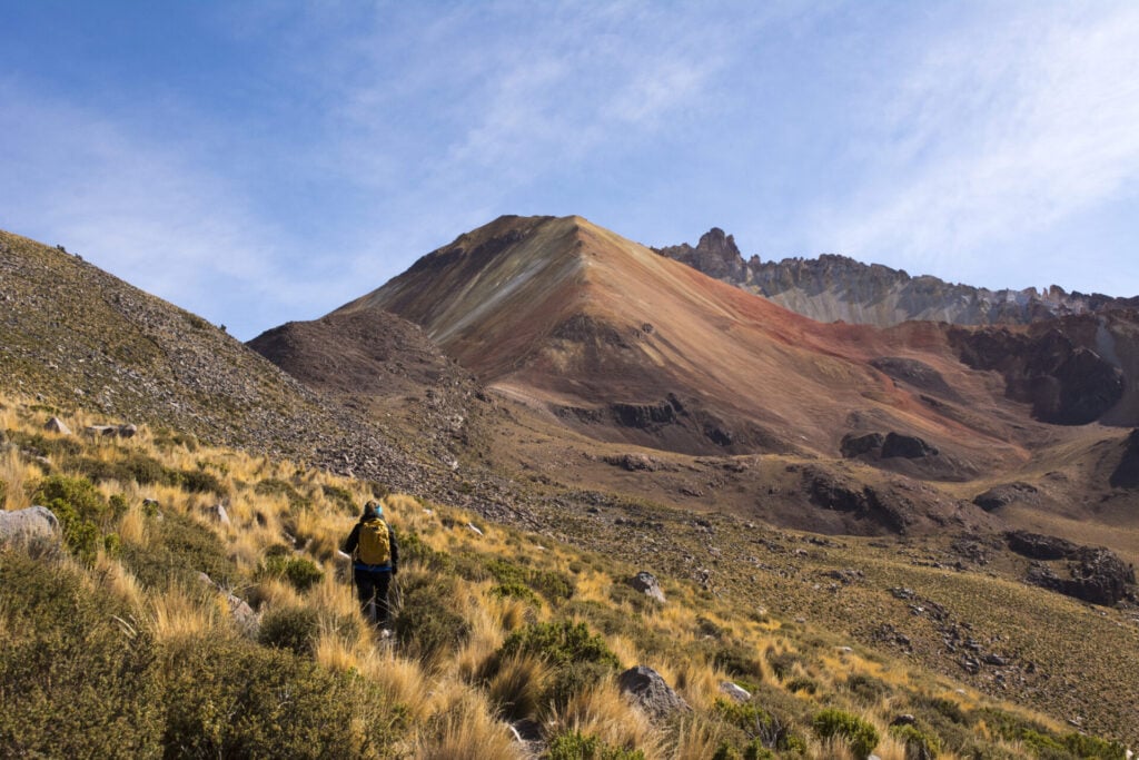 Une personne munie d'un sac à dos jaune marche sur une pente herbeuse et rocheuse en direction d'une montagne multicolore, sous un ciel bleu parsemé de nuages. - Karavaniers