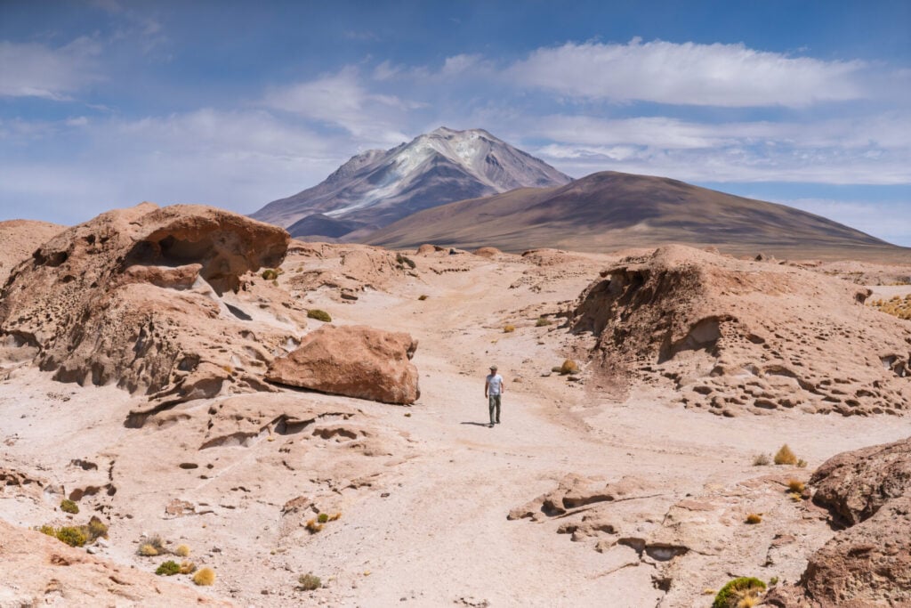 Une personne marche seule dans un paysage désertique et rocailleux, avec une montagne enneigée en arrière-plan, sous un ciel partiellement nuageux. - Karavaniers