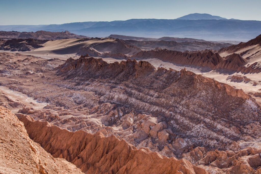 Un vaste paysage désertique et rocailleux avec des crêtes, un terrain brun rougeâtre typique de la région du Sud Lipez et des montagnes lointaines sous un ciel bleu clair. La scène semble sèche et aride, sans végétation visible. - Karavaniers