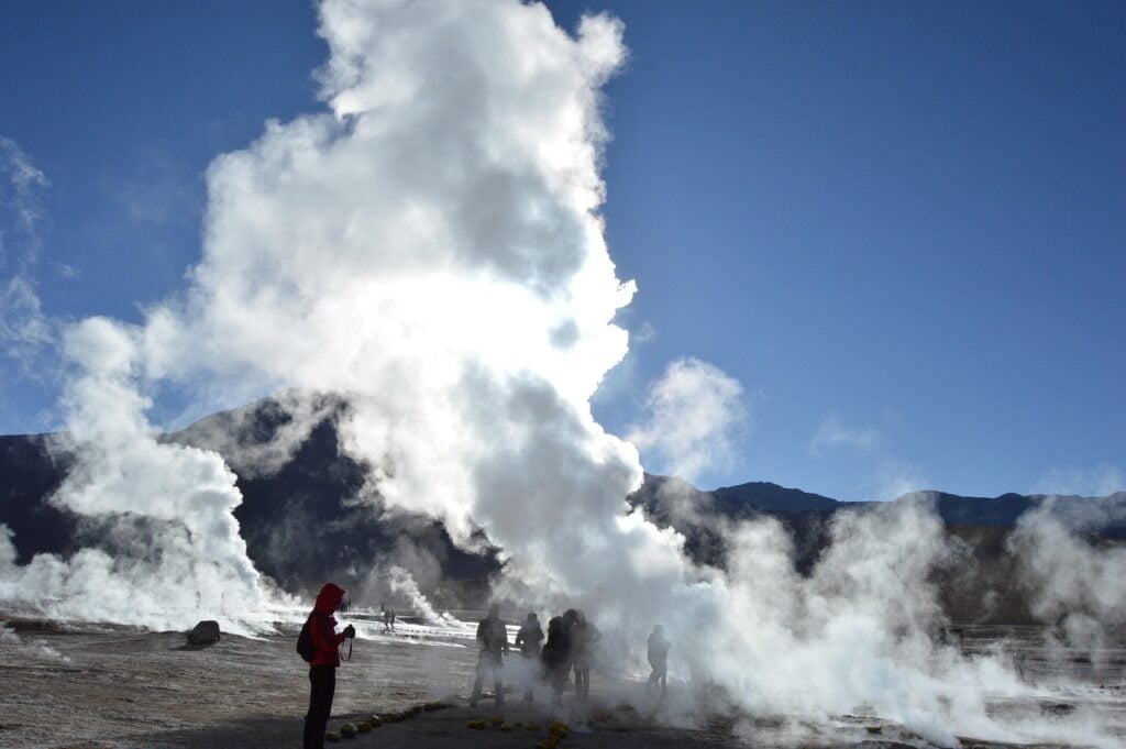 Un groupe de personnes se tient près de geysers ou de fumerolles dans une zone ouverte et rocheuse, avec des montagnes en arrière-plan et un soleil brillant filtrant à travers des nuages de vapeur. - Karavaniers