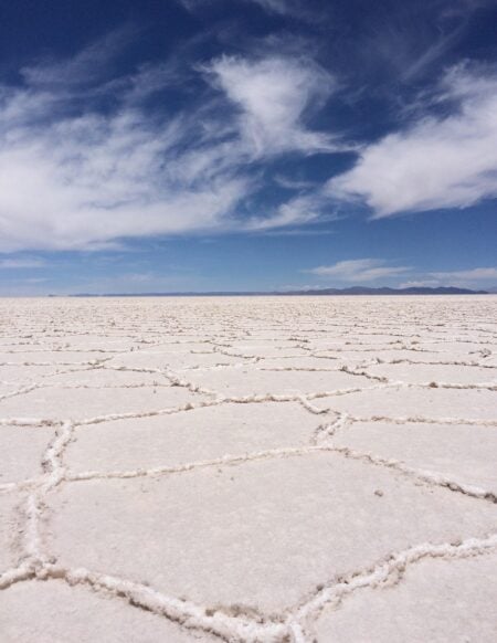 Une étendue plate de croûte de sel blanche forme un motif géométrique sur le sol sous un ciel bleu partiellement nuageux, avec des montagnes faiblement visibles au loin. - Karavaniers