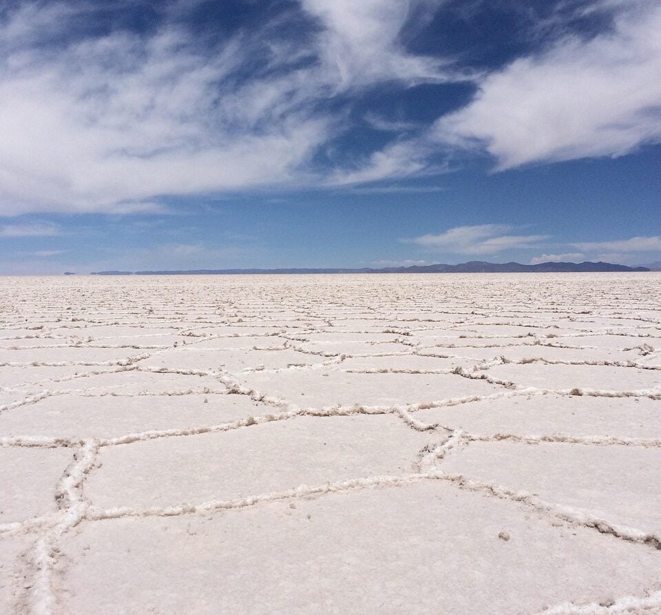 Une étendue plate de croûte de sel blanche forme un motif géométrique sur le sol sous un ciel bleu partiellement nuageux, avec des montagnes faiblement visibles au loin. - Karavaniers