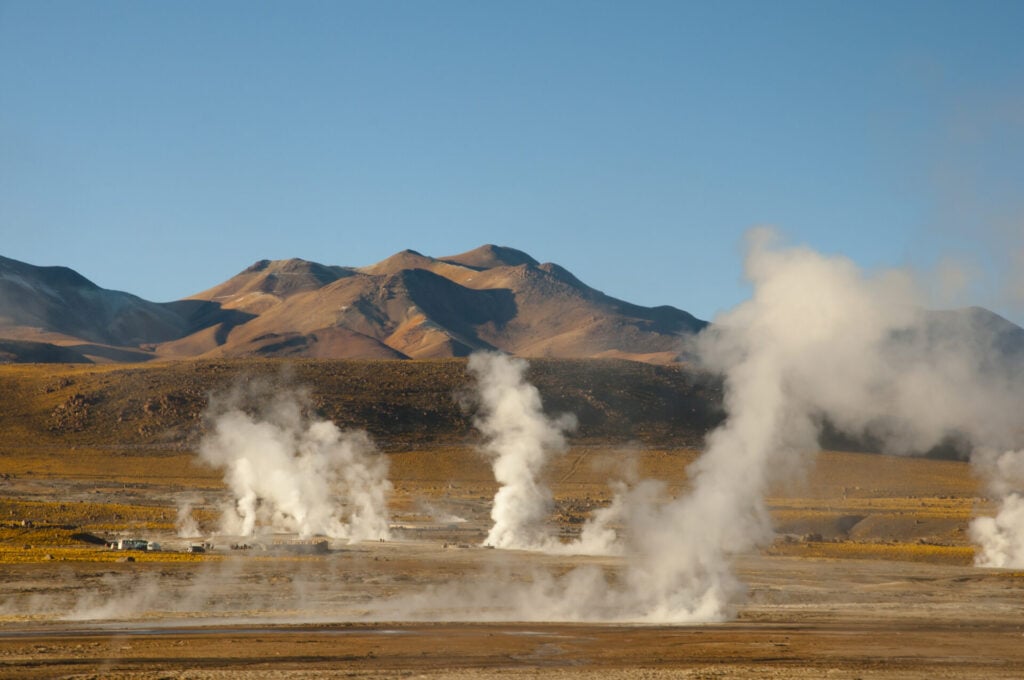 Un paysage aride dans le Sud Lipez avec plusieurs colonnes de vapeur s'élevant des terres de feu, entourées de montagnes sous un ciel bleu clair. - Karavaniers