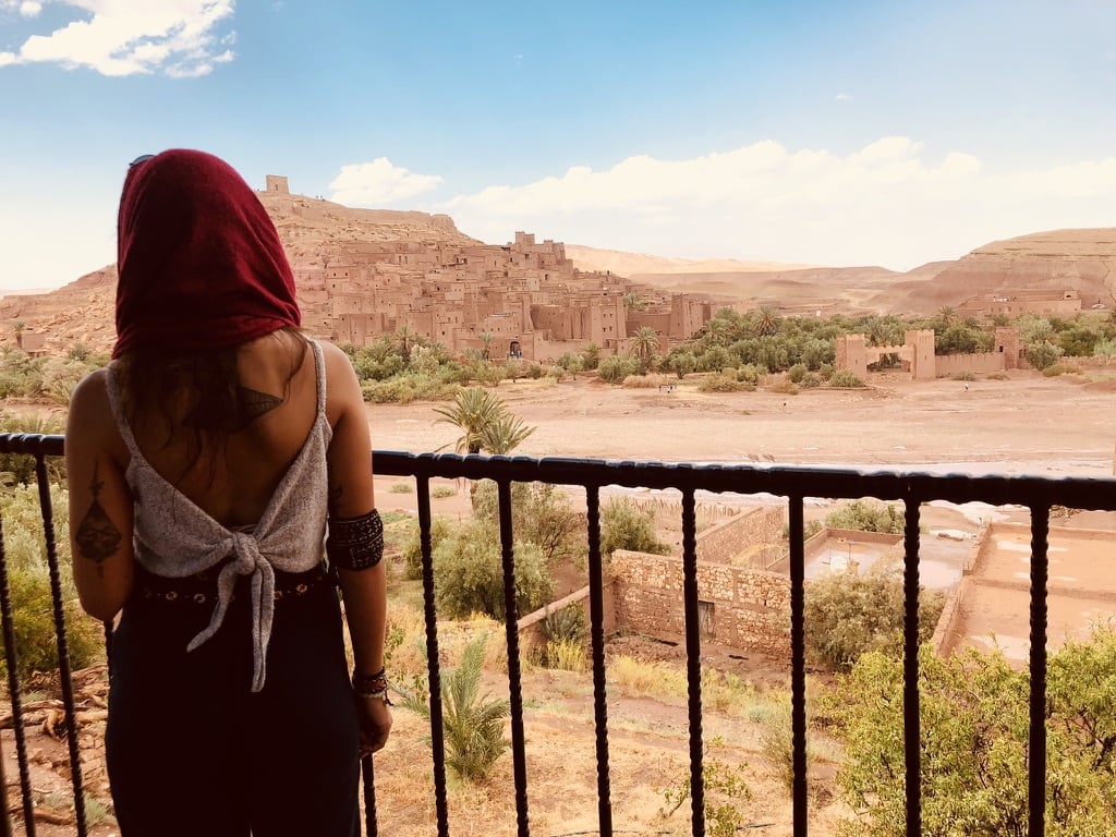 Une personne portant un foulard rouge et un haut sans manches se tient derrière une balustrade noire, regardant un ancien village du désert - comme Mathilde Godbout pourrait le faire - où des bâtiments en briques de boue et une forteresse couronnent la colline sous un ciel partiellement nuageux. - Karavaniers