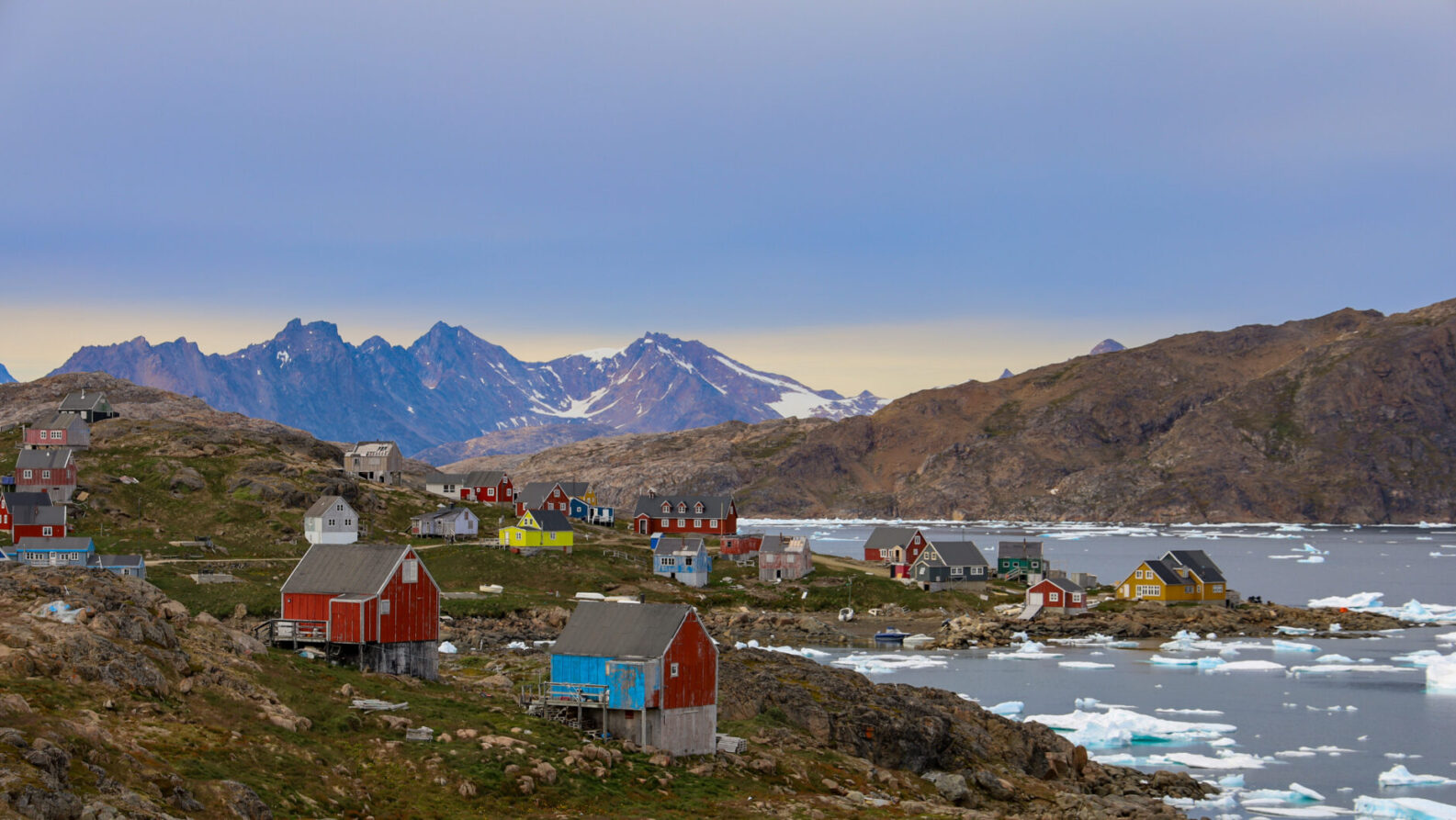 Un petit village côtier du Groenland aux maisons colorées se dresse sur un terrain rocheux au bord de la mer, entouré de glace flottante et de montagnes en arrière-plan, sous un ciel nuageux. - Karavaniers