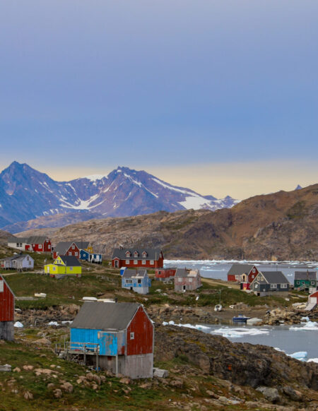 Un petit village côtier du Groenland aux maisons colorées se dresse sur un terrain rocheux au bord de la mer, entouré de glace flottante et de montagnes en arrière-plan, sous un ciel nuageux. - Karavaniers