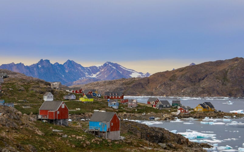 Un petit village côtier du Groenland aux maisons colorées se dresse sur un terrain rocheux au bord de la mer, entouré de glace flottante et de montagnes en arrière-plan, sous un ciel nuageux. - Karavaniers