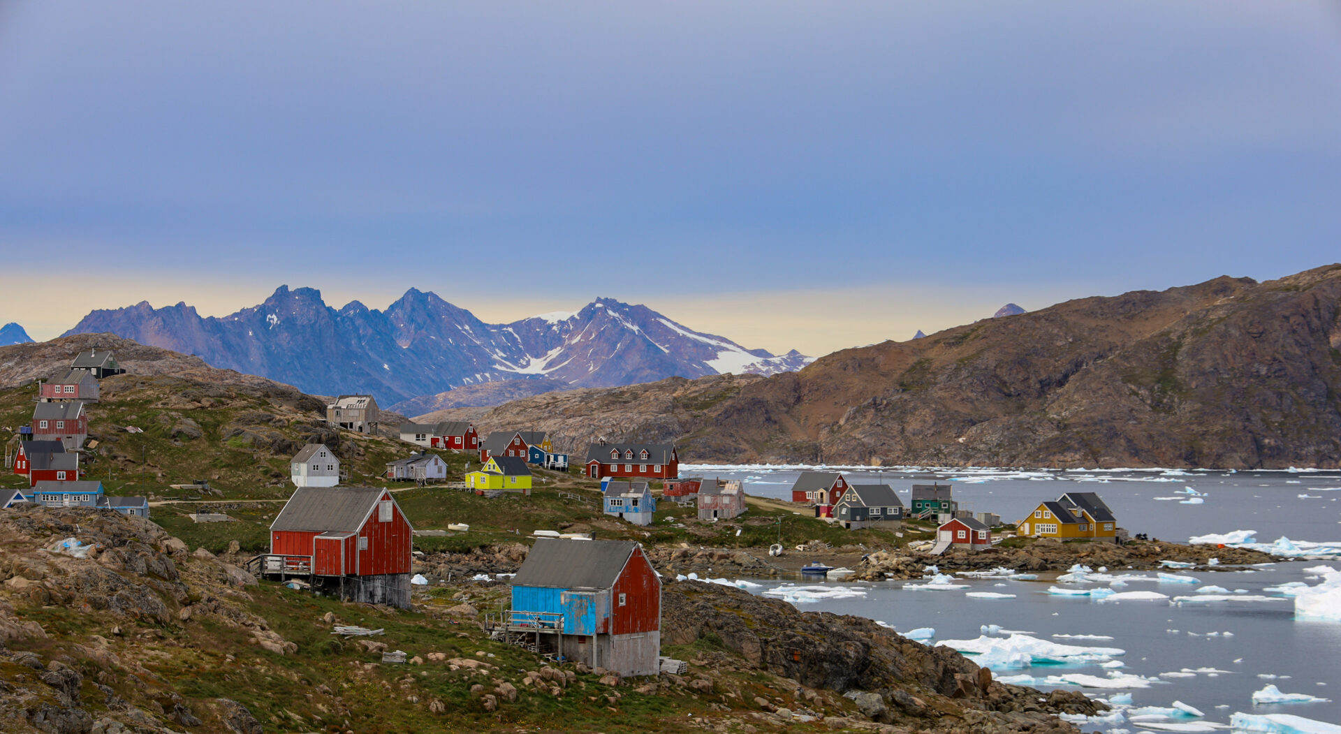 Un petit village côtier du Groenland aux maisons colorées se dresse sur un terrain rocheux au bord de la mer, entouré de glace flottante et de montagnes en arrière-plan, sous un ciel nuageux. - Karavaniers