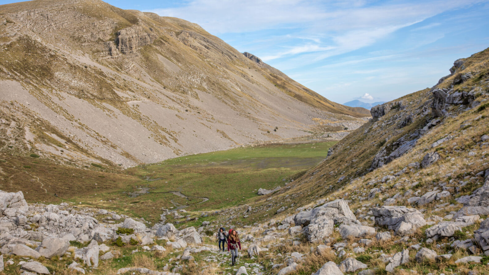 Deux personnes avec des sacs à dos et un chien marchent sur un sentier rocailleux dans une large vallée de montagne, rappelant un trek en Grèce, avec des pentes herbeuses et rocailleuses sous un ciel bleu parsemé de nuages épars. - Karavaniers