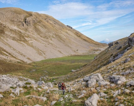 Deux personnes avec des sacs à dos et un chien marchent sur un sentier rocailleux dans une large vallée de montagne, rappelant un trek en Grèce, avec des pentes herbeuses et rocailleuses sous un ciel bleu parsemé de nuages épars. - Karavaniers