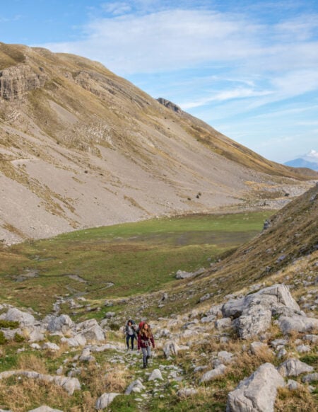 Deux personnes avec des sacs à dos et un chien marchent sur un sentier rocailleux dans une large vallée de montagne, rappelant un trek en Grèce, avec des pentes herbeuses et rocailleuses sous un ciel bleu parsemé de nuages épars. - Karavaniers