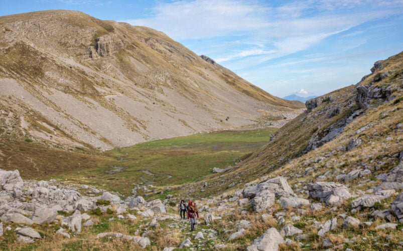 Deux personnes avec des sacs à dos et un chien marchent sur un sentier rocailleux dans une large vallée de montagne, rappelant un trek en Grèce, avec des pentes herbeuses et rocailleuses sous un ciel bleu parsemé de nuages épars. - Karavaniers
