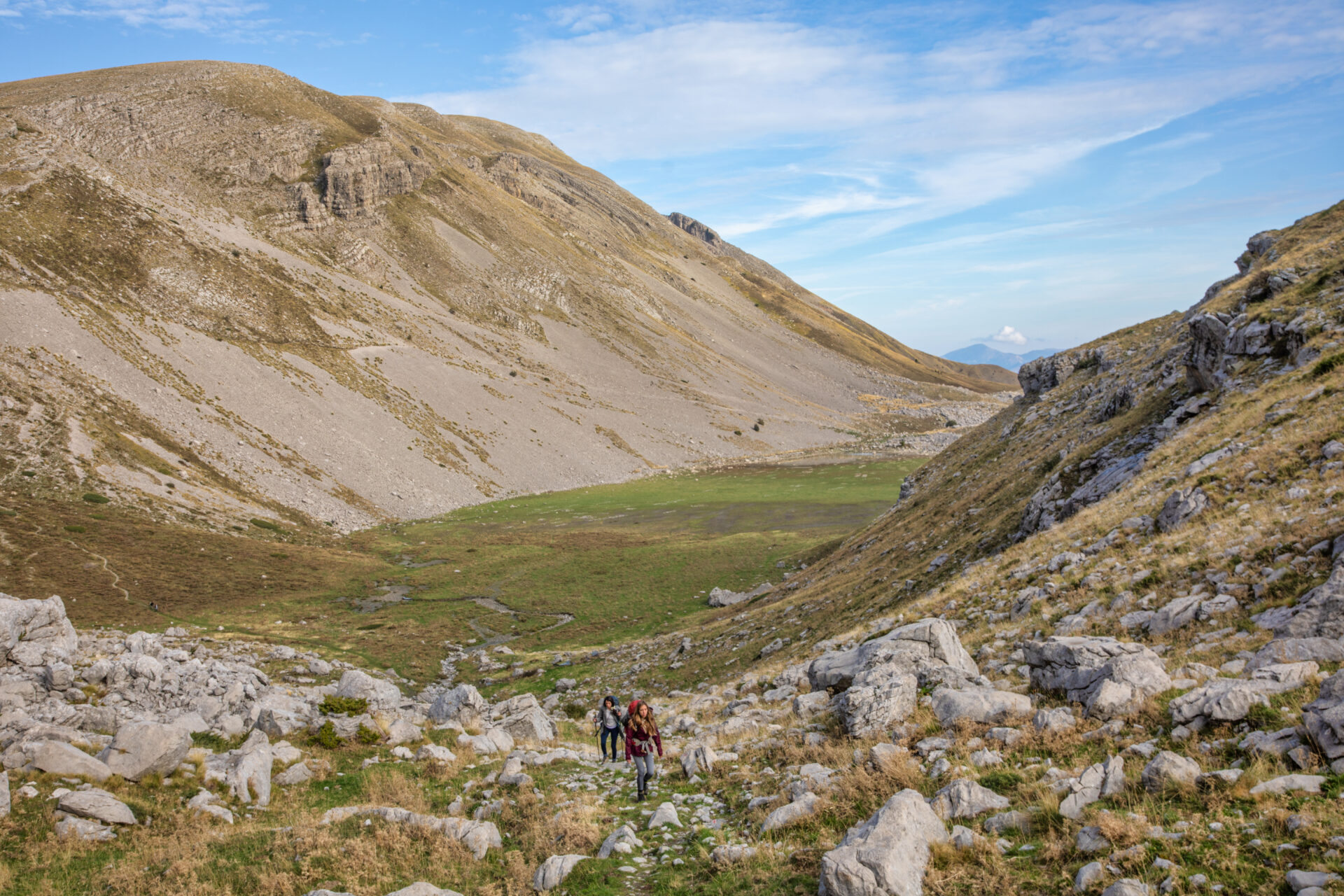 Deux personnes avec des sacs à dos et un chien marchent sur un sentier rocailleux dans une large vallée de montagne, rappelant un trek en Grèce, avec des pentes herbeuses et rocailleuses sous un ciel bleu parsemé de nuages épars. - Karavaniers
