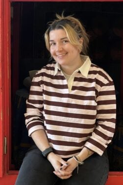 Une personne aux cheveux châtains clairs en queue de cheval, portant une chemise à rayures marron et blanches, un pantalon noir et une montre, est assise à une fenêtre encadrée par des panneaux rouges aux motifs colorés, et sourit à la caméra. - Karavaniers