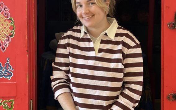 Une personne aux cheveux châtains clairs en queue de cheval, portant une chemise à rayures marron et blanches, un pantalon noir et une montre, est assise à une fenêtre encadrée par des panneaux rouges aux motifs colorés, et sourit à la caméra. - Karavaniers