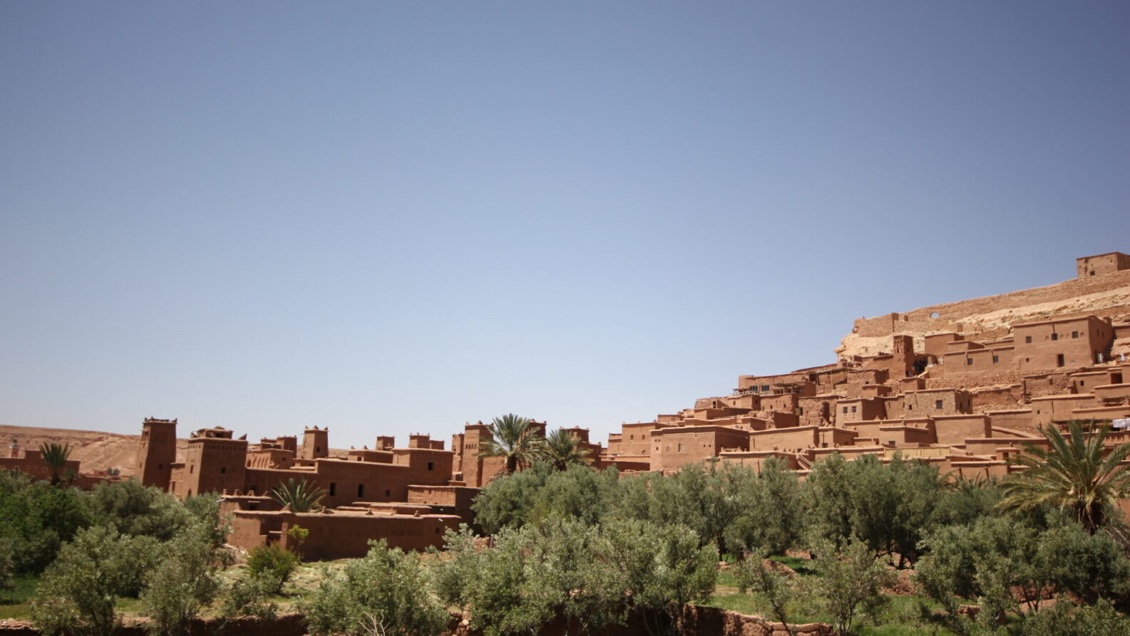 Un groupe de bâtiments traditionnels en terre sur le flanc d'une colline sous un ciel bleu clair, avec des arbres verts et de la végétation au premier plan - une scène pittoresque souvent admirée lors des randonnées au Maroc. - Karavaniers