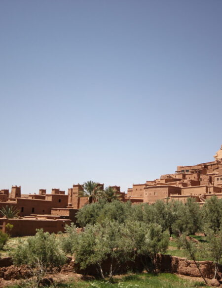 Un groupe de bâtiments traditionnels en terre sur le flanc d'une colline sous un ciel bleu clair, avec des arbres verts et de la végétation au premier plan - une scène pittoresque souvent admirée lors des randonnées au Maroc. - Karavaniers