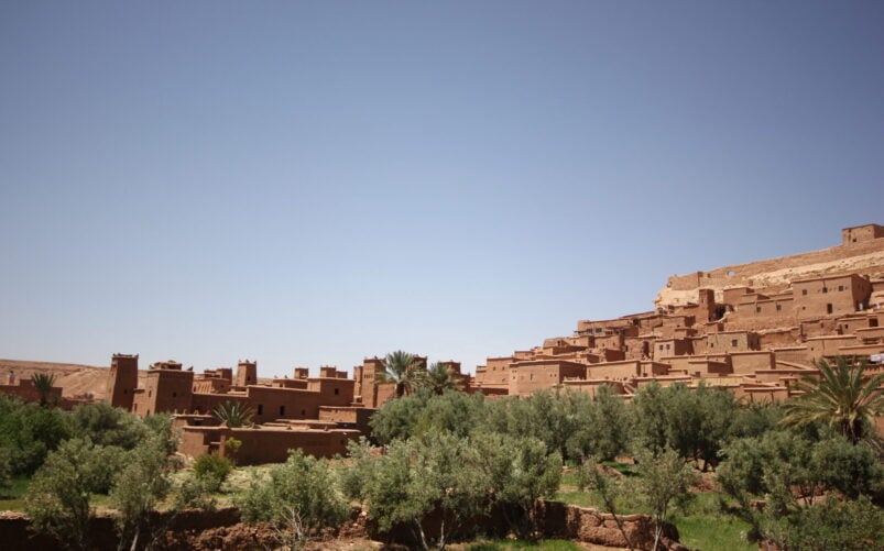 Un groupe de bâtiments traditionnels en terre sur le flanc d'une colline sous un ciel bleu clair, avec des arbres verts et de la végétation au premier plan - une scène pittoresque souvent admirée lors des randonnées au Maroc. - Karavaniers
