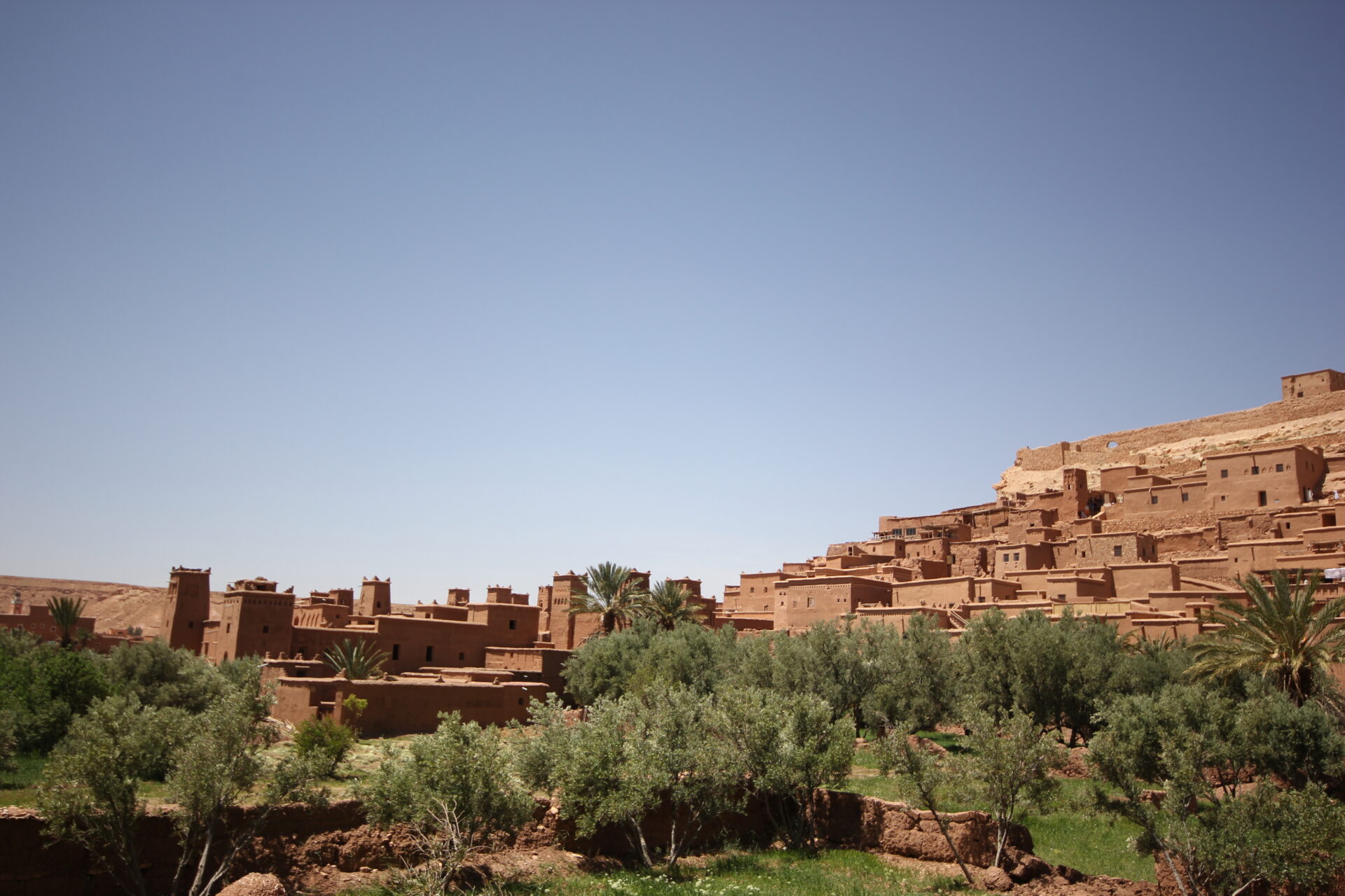 Un groupe de bâtiments traditionnels en terre sur le flanc d'une colline sous un ciel bleu clair, avec des arbres verts et de la végétation au premier plan - une scène pittoresque souvent admirée lors des randonnées au Maroc. - Karavaniers