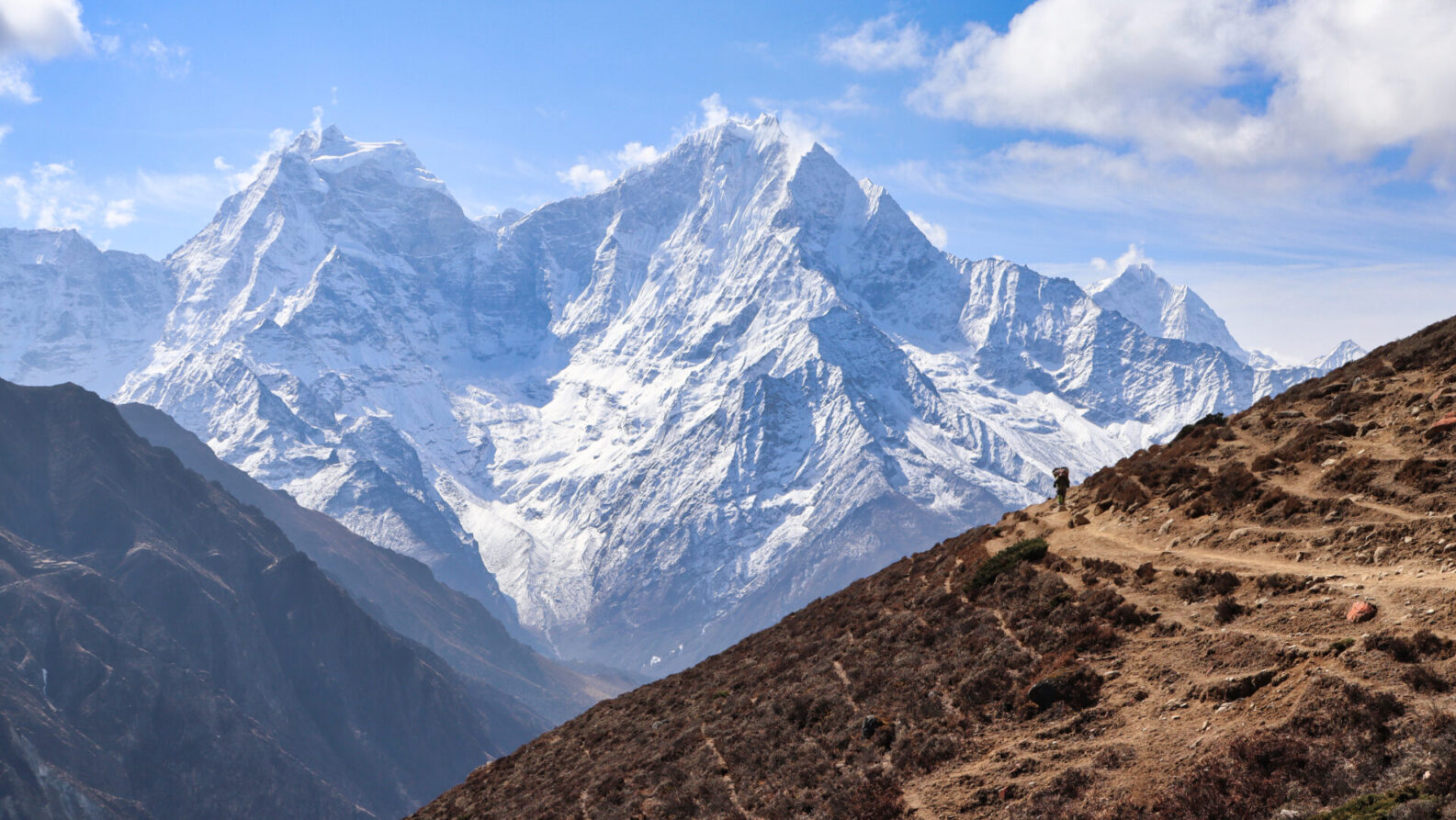 Une personne marche sur un sentier de terre à flanc de colline au Ladakh, avec des montagnes enneigées en arrière-plan, sous un ciel partiellement nuageux. - Karavaniers