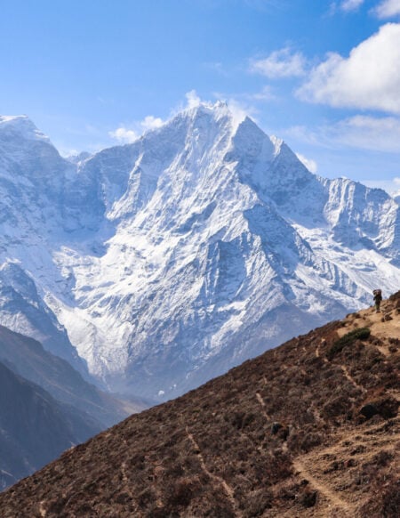 Une personne marche sur un sentier de terre à flanc de colline au Ladakh, avec des montagnes enneigées en arrière-plan, sous un ciel partiellement nuageux. - Karavaniers