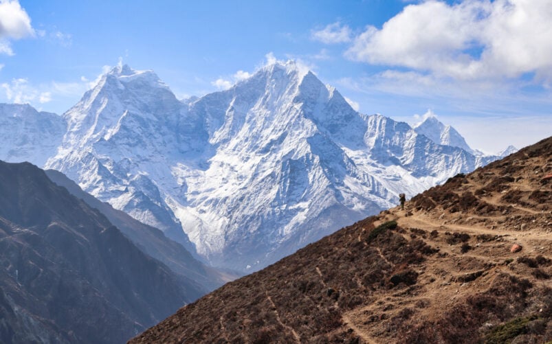 Une personne marche sur un sentier de terre à flanc de colline au Ladakh, avec des montagnes enneigées en arrière-plan, sous un ciel partiellement nuageux. - Karavaniers
