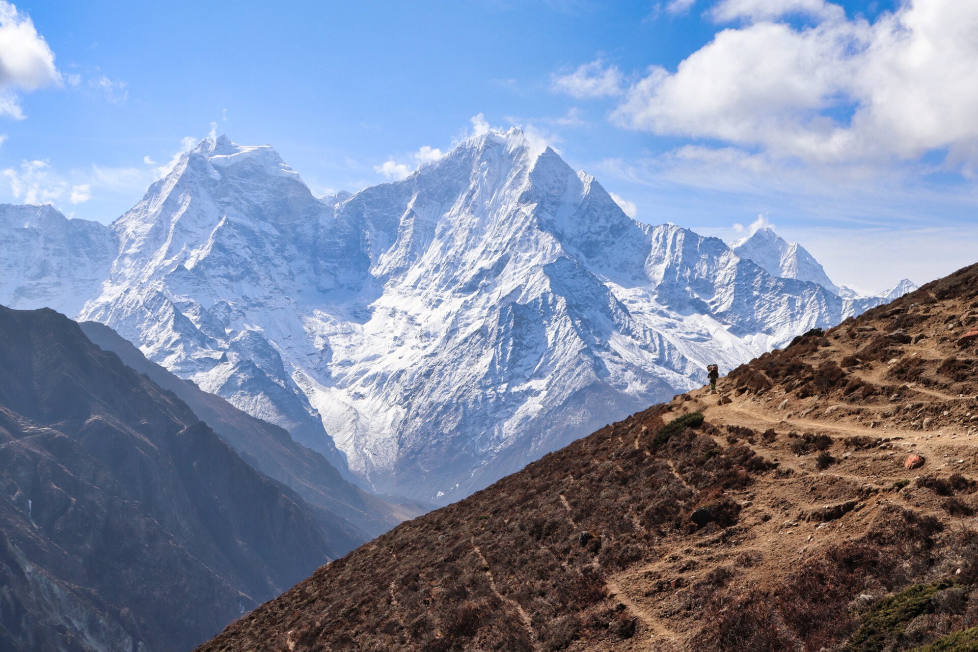 Une personne marche sur un sentier de terre à flanc de colline au Ladakh, avec des montagnes enneigées en arrière-plan, sous un ciel partiellement nuageux. - Karavaniers