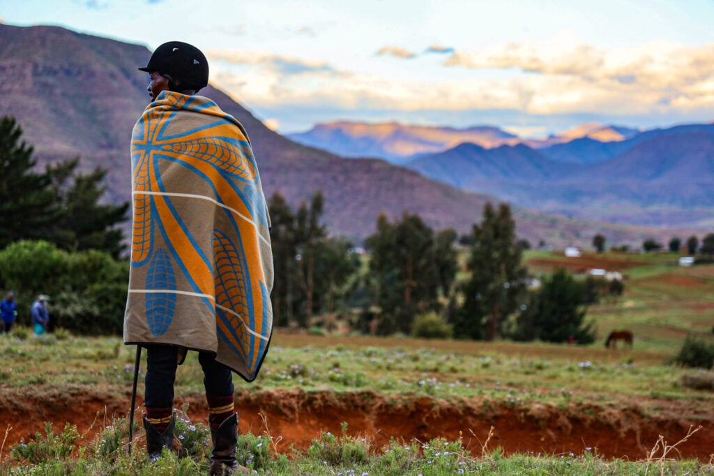 Une personne portant une couverture et un chapeau à motifs se tient face à une vallée pittoresque en Afrique du Sud, comme si elle faisait une pause au cours d'une randonnée. Des montagnes s'élèvent à l'arrière-plan sous un ciel partiellement nuageux, avec des arbres, de l'herbe et des silhouettes lointaines dans le paysage. - Karavaniers
