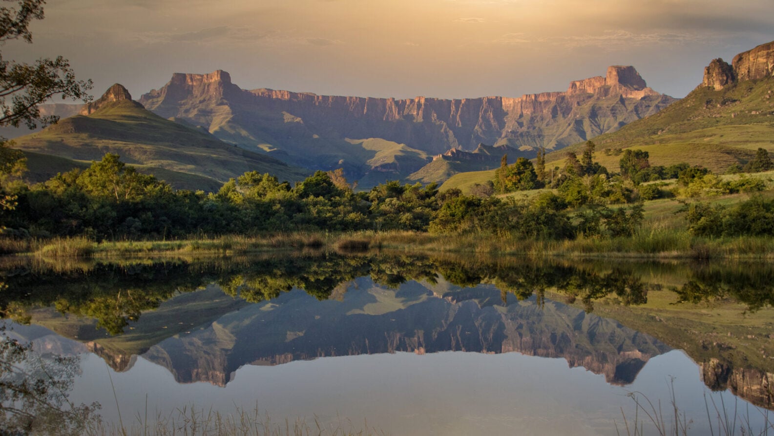 Une chaîne de montagnes aux sommets plats se reflète dans une étendue d'eau calme, entourée de collines vertes et de végétation, sous un ciel nuageux au coucher du soleil. - Karavaniers