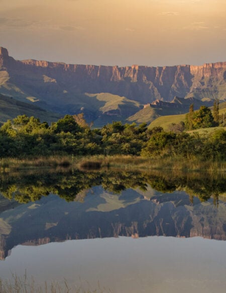Une chaîne de montagnes aux sommets plats se reflète dans une étendue d'eau calme, entourée de collines vertes et de végétation, sous un ciel nuageux au coucher du soleil. - Karavaniers