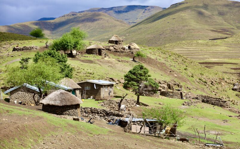 Plusieurs petites huttes en pierre et au toit de chaume sont dispersées dans un paysage vallonné et herbeux, avec quelques arbres et clôtures, sous un ciel partiellement nuageux. Des collines et des montagnes sont visibles à l'arrière-plan. - Karavaniers