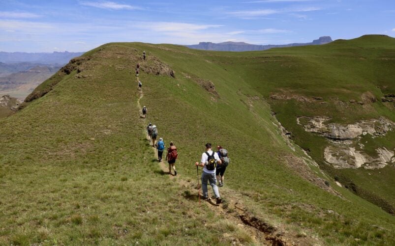 Un groupe de randonneurs marche en ligne le long d'un sentier étroit sur une crête herbeuse, entourée de collines ondulantes et de montagnes lointaines sous un ciel bleu. - Karavaniers